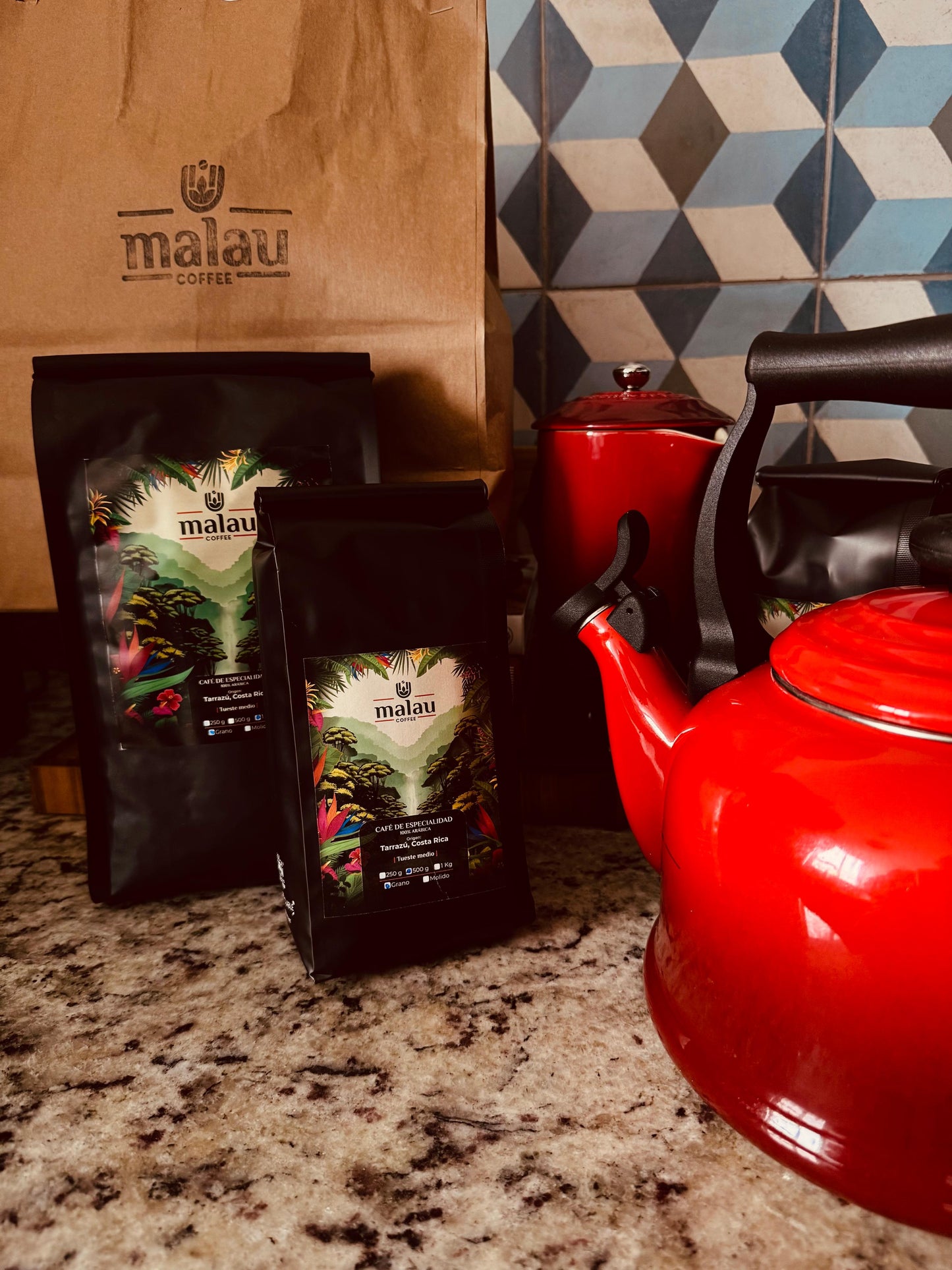 Dos bolsas de Malau Coffee junto a dos cafeteras rojas sobre una encimera de mármol.