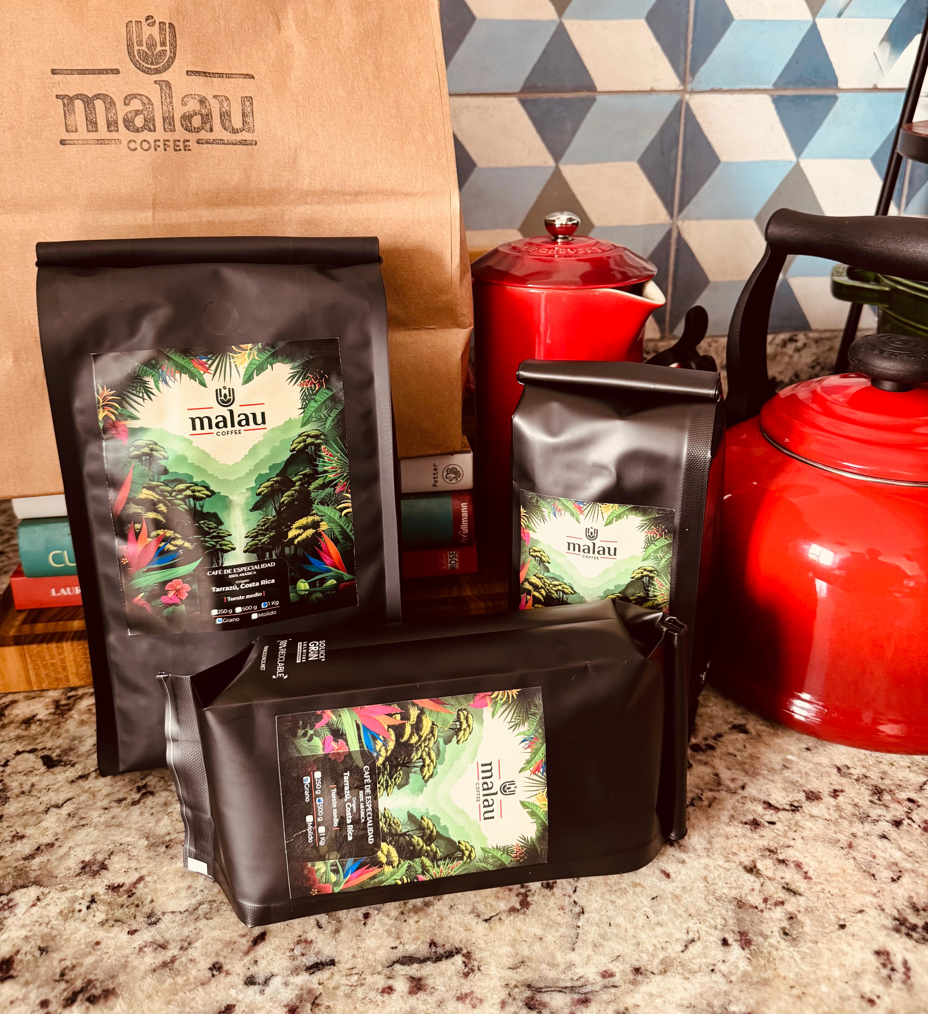 3 bolsas de Malau Coffee en cocina con cafeteras rojas y libros en el fondo.