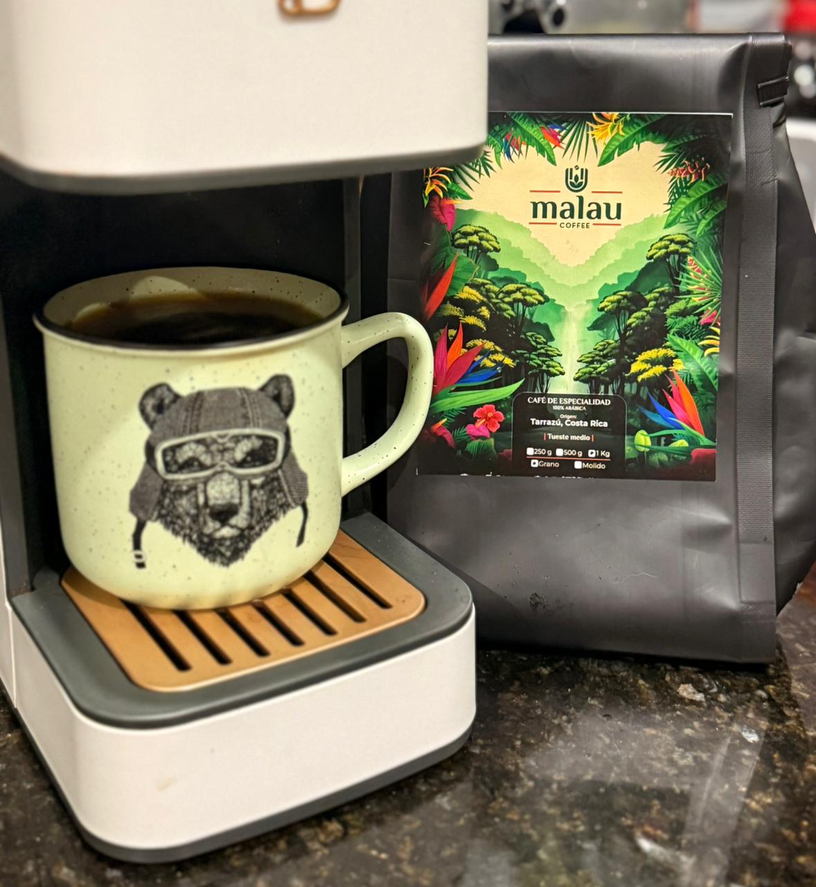 Taza de café con cara de animal y gorro de piloto, junto a una cafetera con café y una bolsa de Malau Coffee.